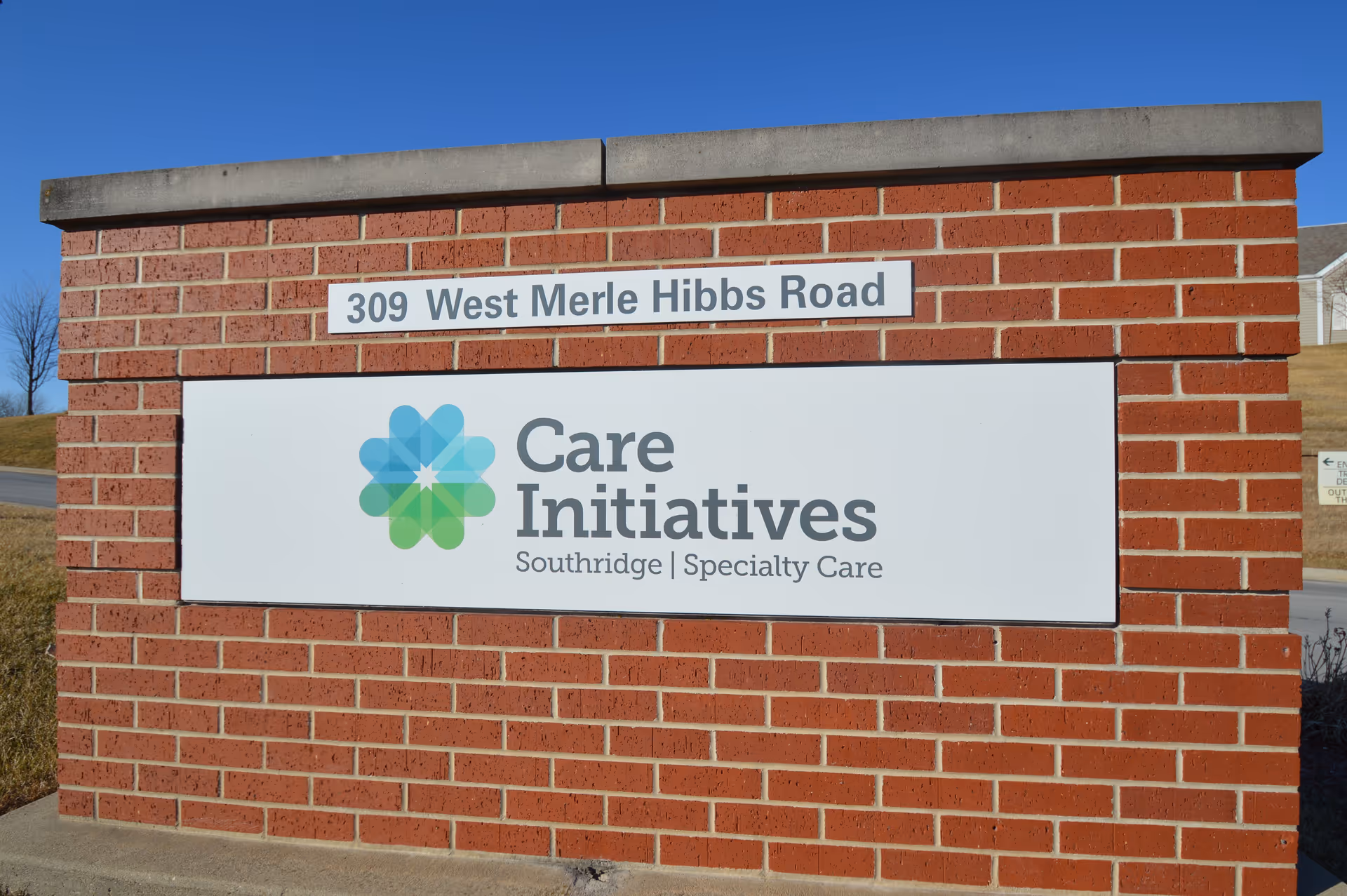 A brick signpost with a white sign displaying the address '309 West Merle Hibbs Road' and the logo and name 'Care Initiatives Southridge Specialty Care' under a clear blue sky.