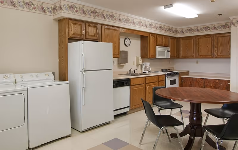 Communal kitchen with white fridge, dishwasher, washer and dryer, oak cabinets, a round wooden table and black chairs.