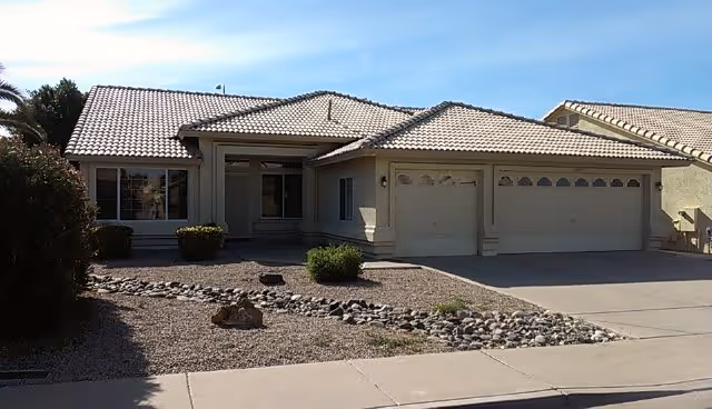 Single-story house with a tiled roof, a three-car garage, and a front yard landscaped with rocks and small bushes under a clear blue sky.