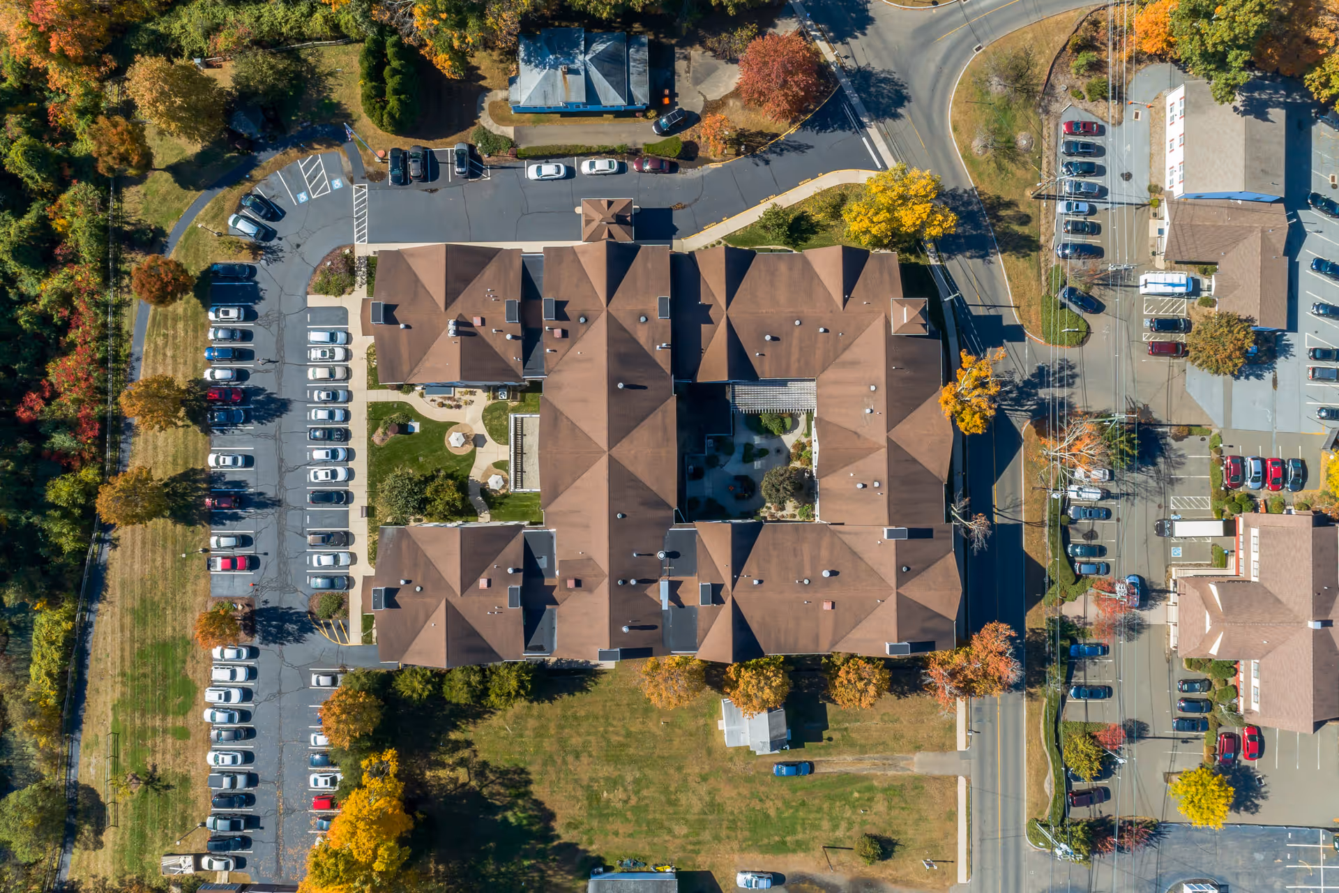 Aerial view of HarborChase of Madison senior living facility showing a large building with a brown roof, surrounded by parking lots filled with cars, trees with autumn foliage, and adjacent roads.