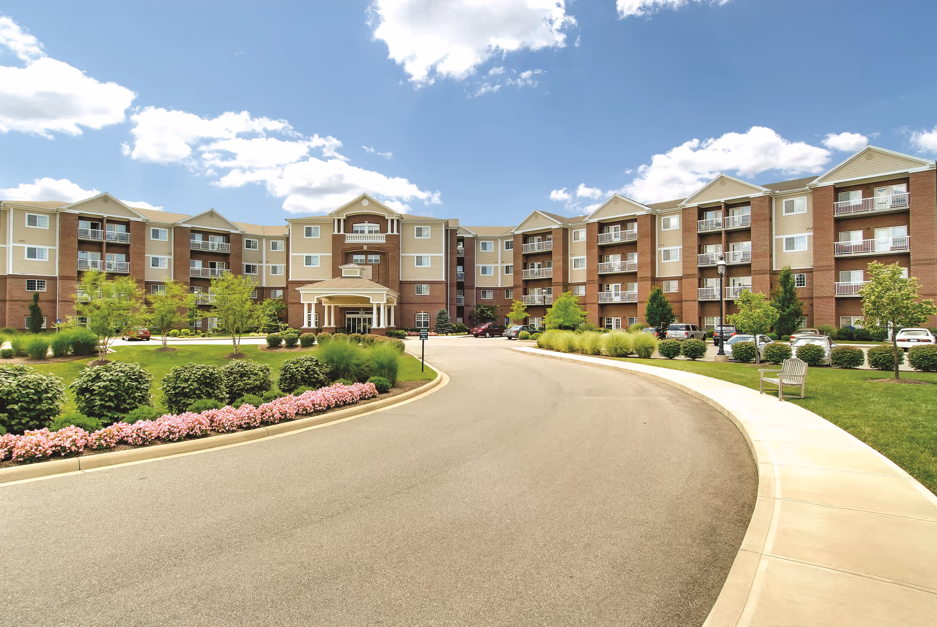 Front exterior view of Barrington of Oakley, a multi-story senior living facility with a curved driveway, landscaped gardens with bushes and flowers, and a clear blue sky with some clouds.