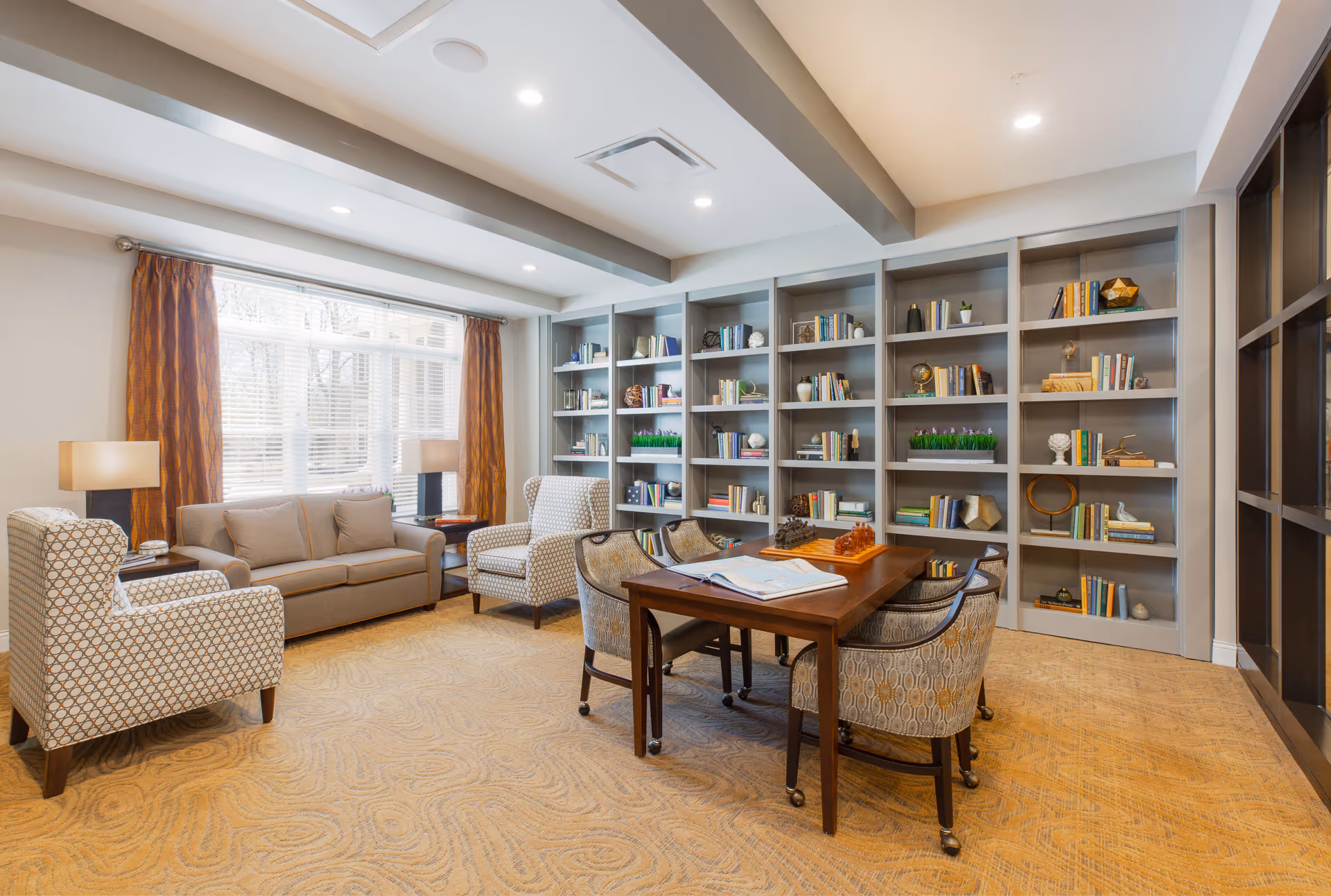 Bright communal lounge with wall-to-wall bookshelves, a central table and upholstered seating by a large window.