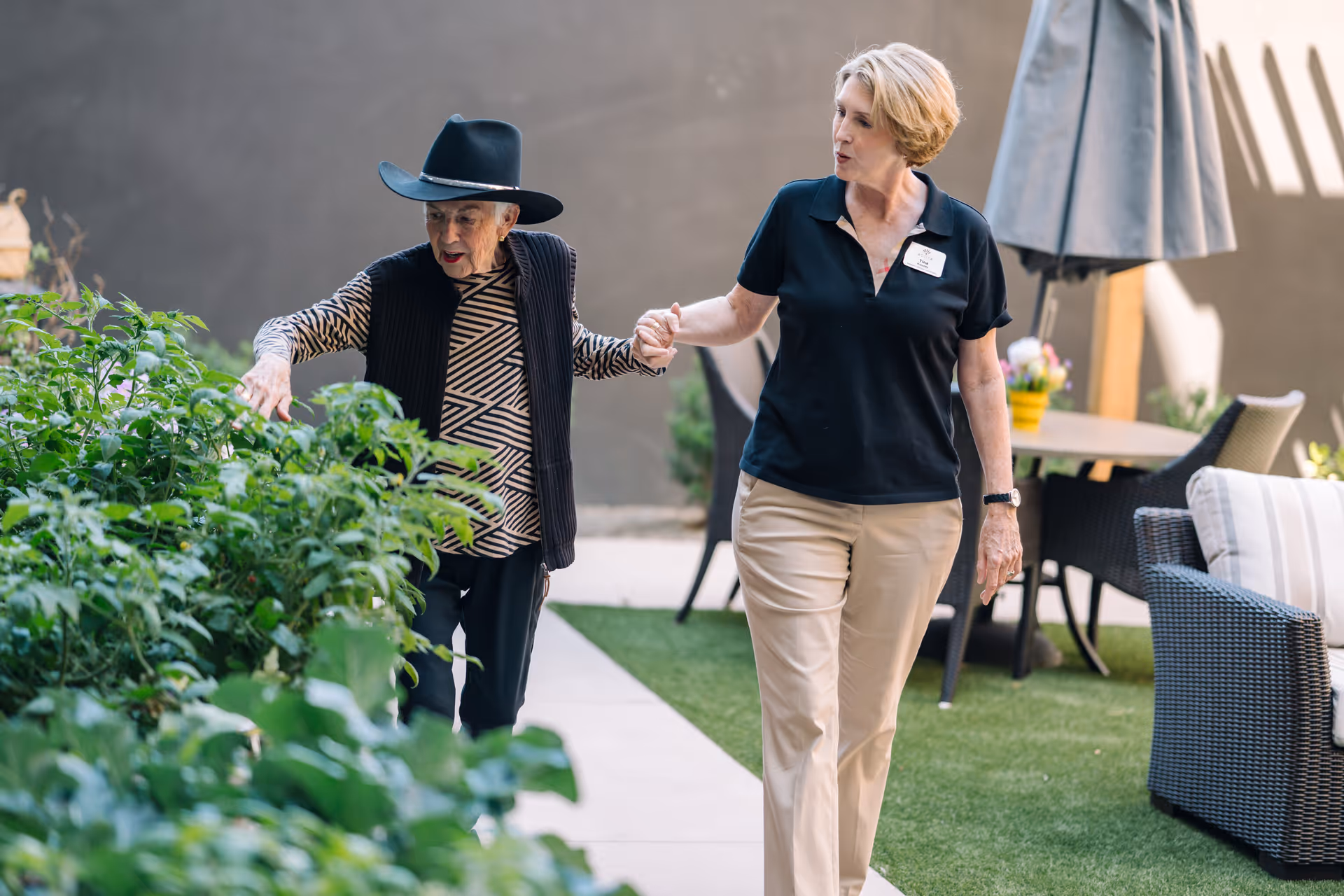 An elderly woman wearing a black hat and patterned shirt is being assisted by a caregiver in a black polo shirt and beige pants as they walk together along a garden path with green plants and outdoor furniture nearby.
