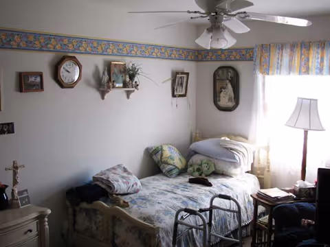 A small bedroom with a single bed covered in floral bedding and pillows, a walker placed at the foot of the bed, a nightstand with a lamp and books, and various wall decorations including a clock, framed pictures, and a small shelf with plants. A ceiling fan with lights is visible above, and a window with floral curtains lets in natural light.