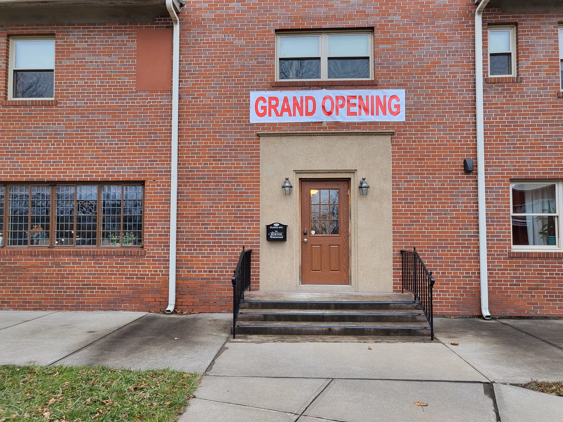 Brick building entrance with steps and a brown door beneath a 'GRAND OPENING' banner.