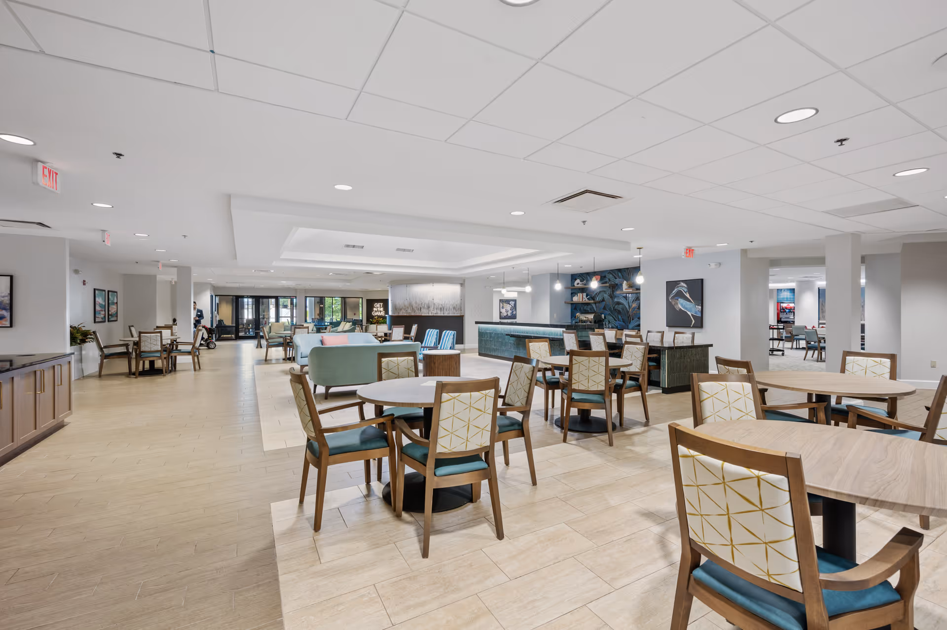 Spacious and modern common area in a senior living facility with multiple round wooden tables and chairs arranged neatly. The room features a seating area with sofas and armchairs, a decorative wall with blue and green leaf patterns, and bright overhead lighting. Large windows and glass doors are visible in the background, allowing natural light to enter.