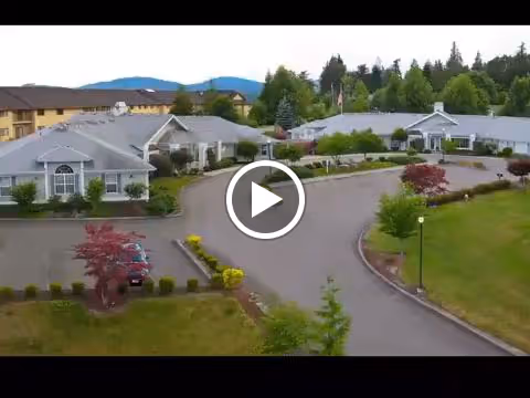 Aerial view of a senior living facility with multiple single-story buildings surrounded by greenery, trees, and a paved driveway. Mountains are visible in the background under a clear sky.