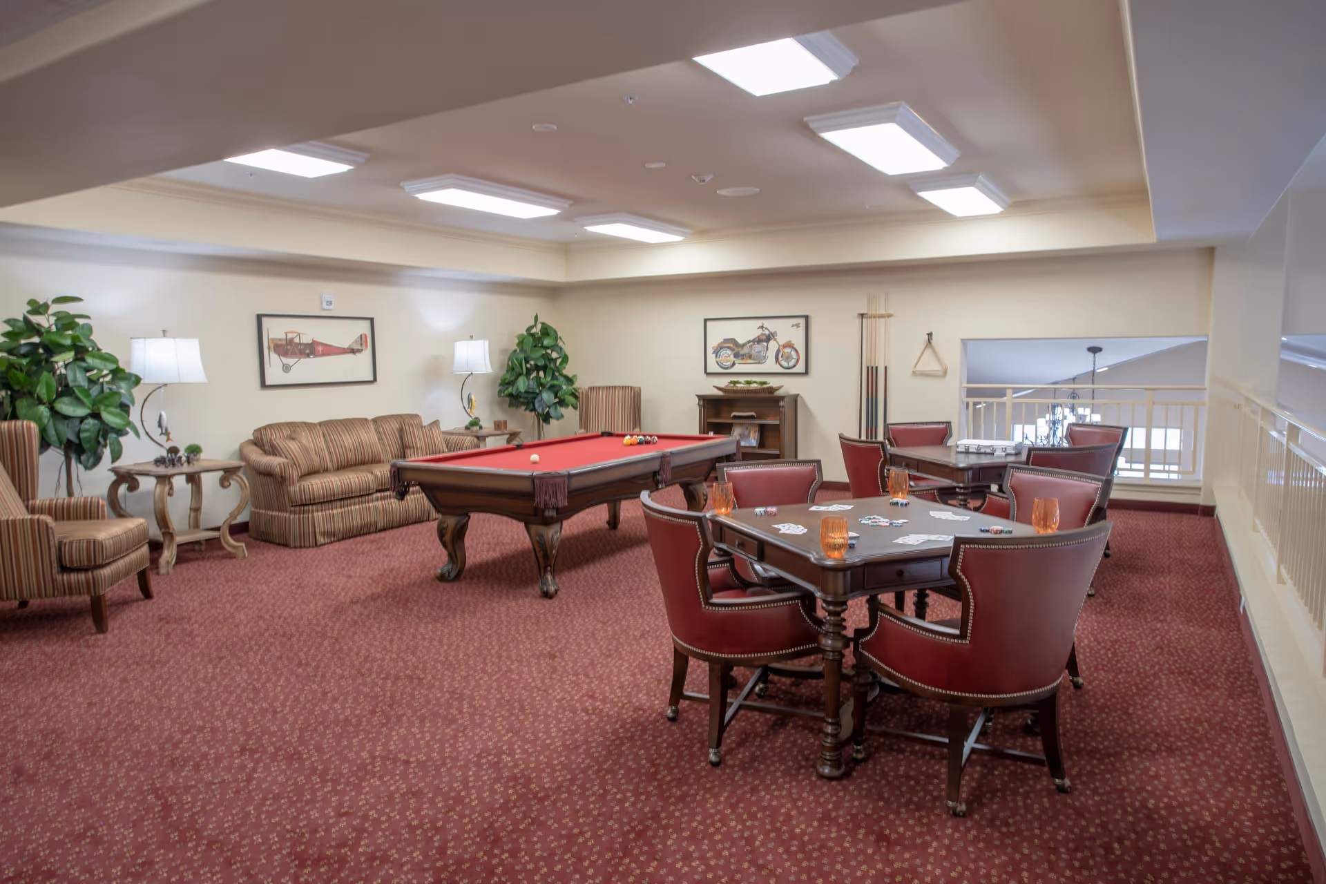A cozy recreational room featuring a red pool table in the center, surrounded by a striped sofa and armchairs. There are two card tables with red leather chairs, each set with playing cards and orange glasses. The room has warm lighting from ceiling fixtures and table lamps, with framed artwork on the walls and green plants adding a touch of nature.
