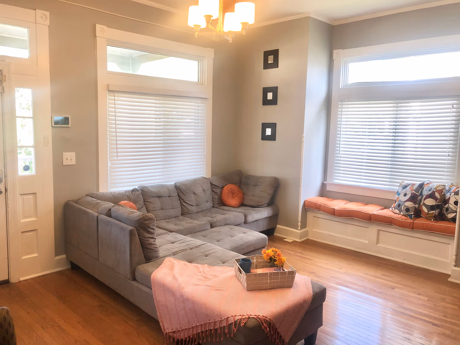A bright living room with a gray sectional sofa adorned with orange round pillows, a pink throw blanket, and a tray with a cup and flowers on the ottoman. The room features hardwood floors, large windows with white blinds, a built-in window seat with orange cushions and patterned pillows, and a modern ceiling light fixture.