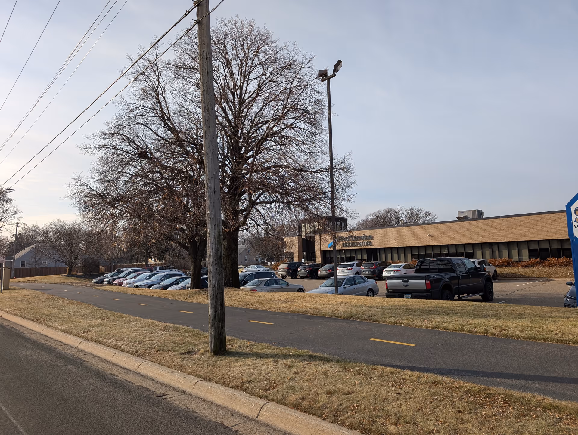 Exterior view of a single-story brick building with a parking lot filled with cars in front. There is a large leafless tree and a streetlight near the parking area. A paved pathway runs parallel to the road in the foreground.