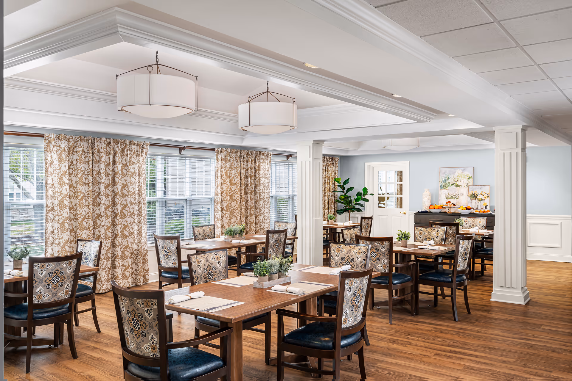 A bright and inviting dining room with wooden tables and patterned chairs arranged neatly. The room features large windows with patterned curtains allowing natural light to fill the space. There are decorative plants on the tables and a sideboard with artwork and additional decor against the far wall. The ceiling has recessed lighting and two large pendant lights.