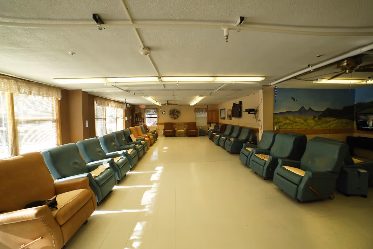 Long communal lounge with rows of reclining chairs lining both sides of a wide room and windows along the left wall.