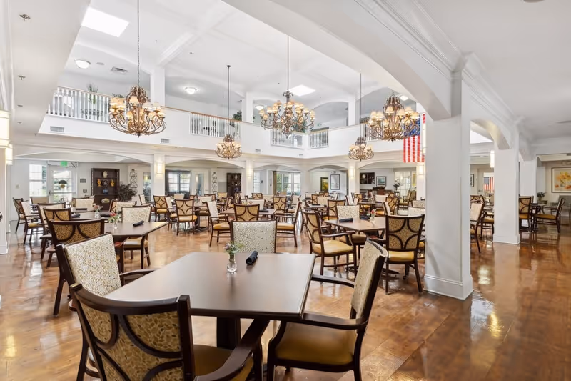 Bright two-story dining room with numerous tables and upholstered chairs under ornate chandeliers.