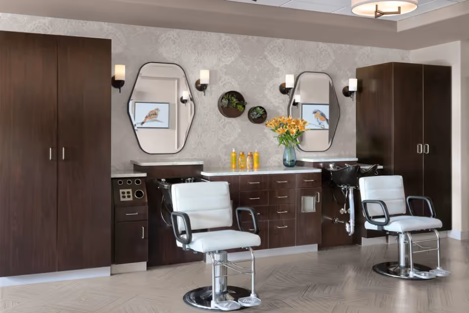 Interior salon area with two white styling chairs, dark wood cabinetry, sinks, mirrors, and a vase of flowers on the counter.
