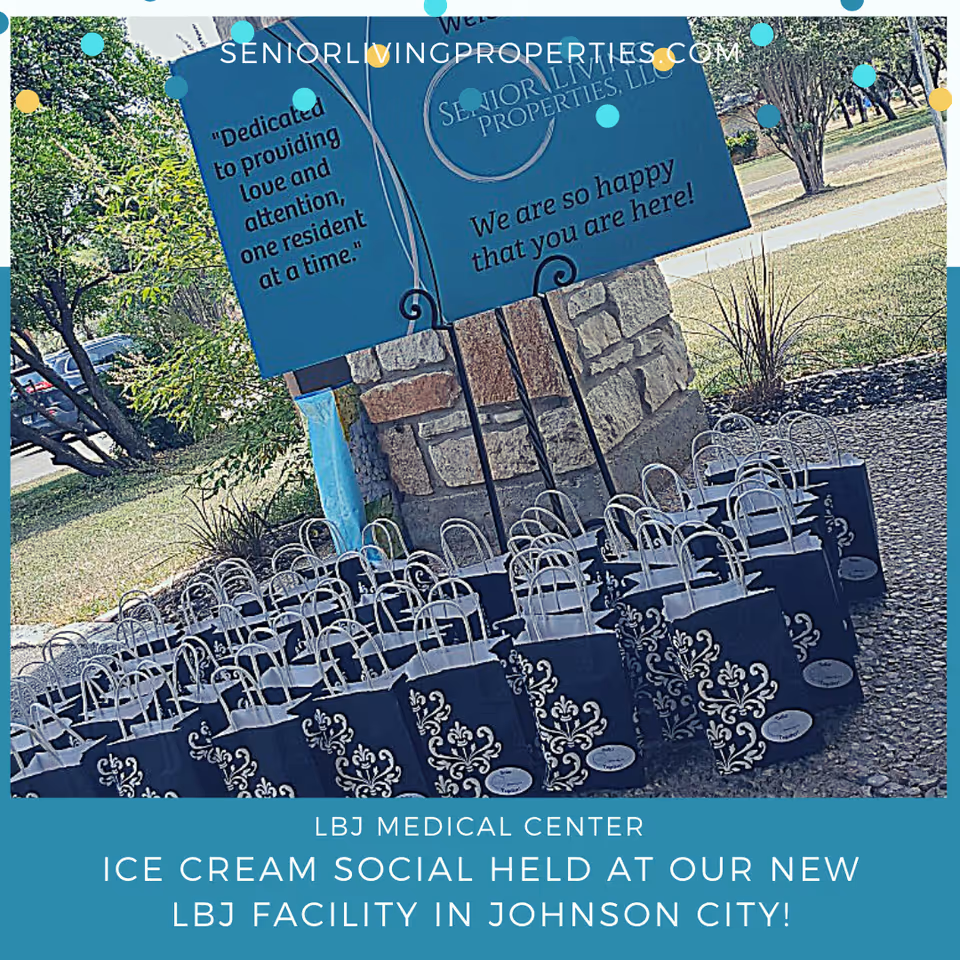 Outdoor scene showing a stone signpost with a blue sign that reads 'Dedicated to providing love and attention, one resident at a time.' and 'We are so happy that you are here!' In front of the sign, there are multiple black gift bags with white decorative patterns and handles arranged on the ground. Trees and grass are visible in the background.