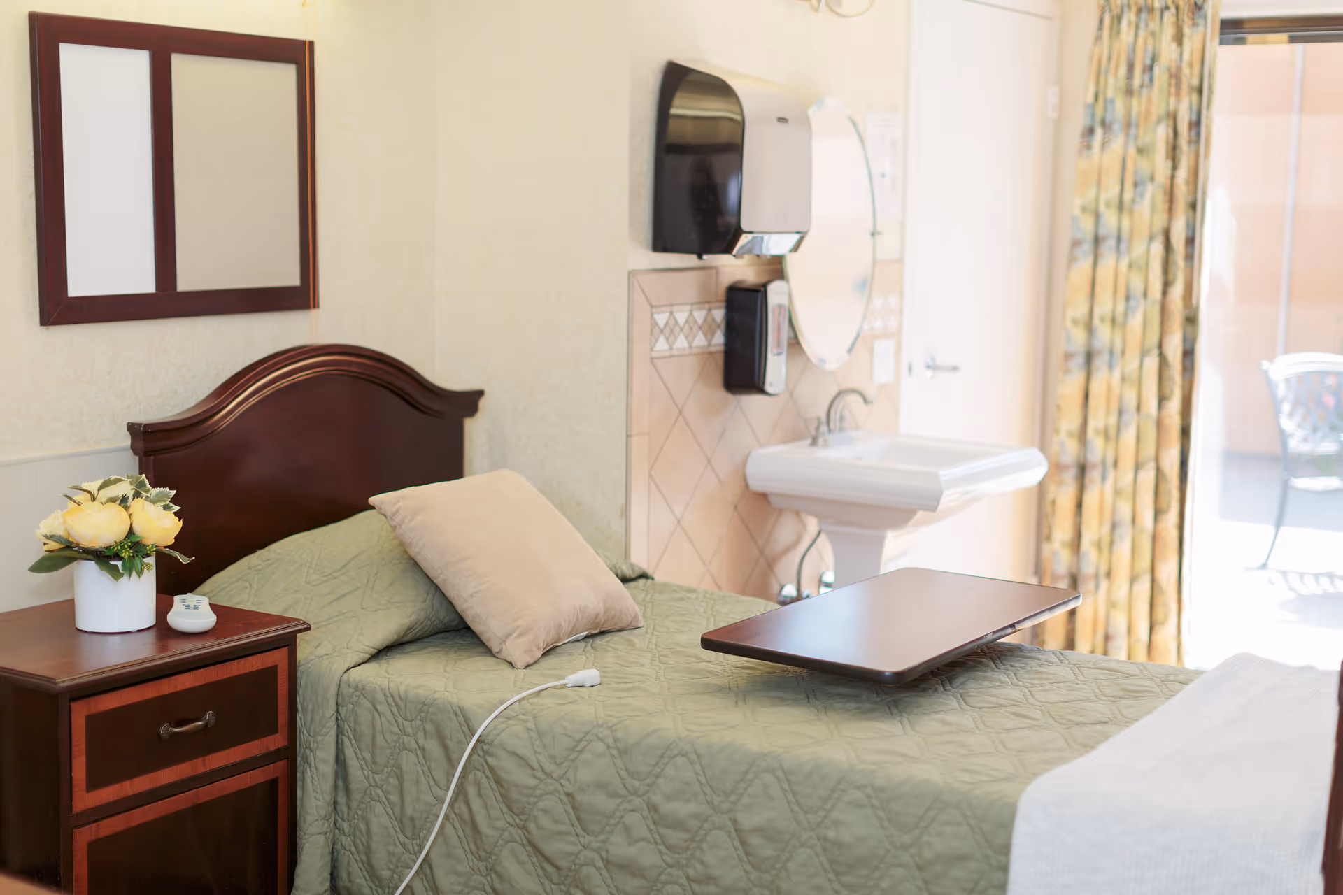 A single bed with a green quilt and beige pillow in a room. Next to the bed is a wooden nightstand with a vase of yellow flowers and a remote control. On the wall above the bed is a framed mirror. A white sink with a round mirror above it and a soap dispenser is mounted on the wall near the bed. A tray table is placed on the bed. A window with floral curtains lets in natural light, and an outdoor chair is visible outside.