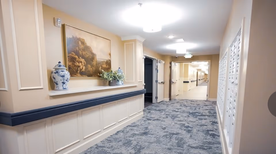 A well-lit hallway in a senior living facility with beige walls, blue-gray carpet, and white trim. The left wall features a shelf with decorative vases and a framed landscape painting. The right wall has a bank of mailboxes. The hallway extends into the distance with open double doors and ceiling lights.