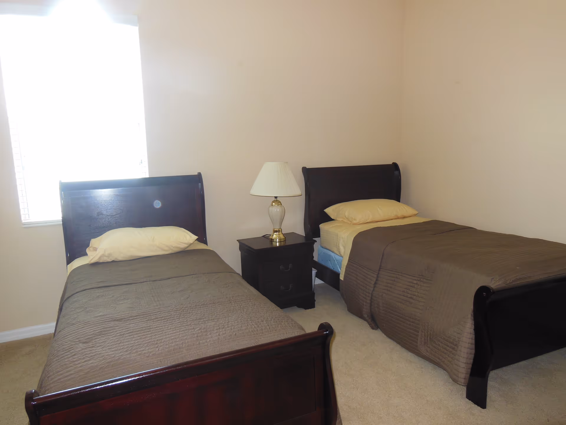 A simple bedroom with two single beds featuring dark wooden frames, beige pillows, and brown bedspreads. A small dark wooden nightstand with a white lamp is positioned between the beds. There is a window with blinds on the left side of the room, and the walls are painted a light beige color.
