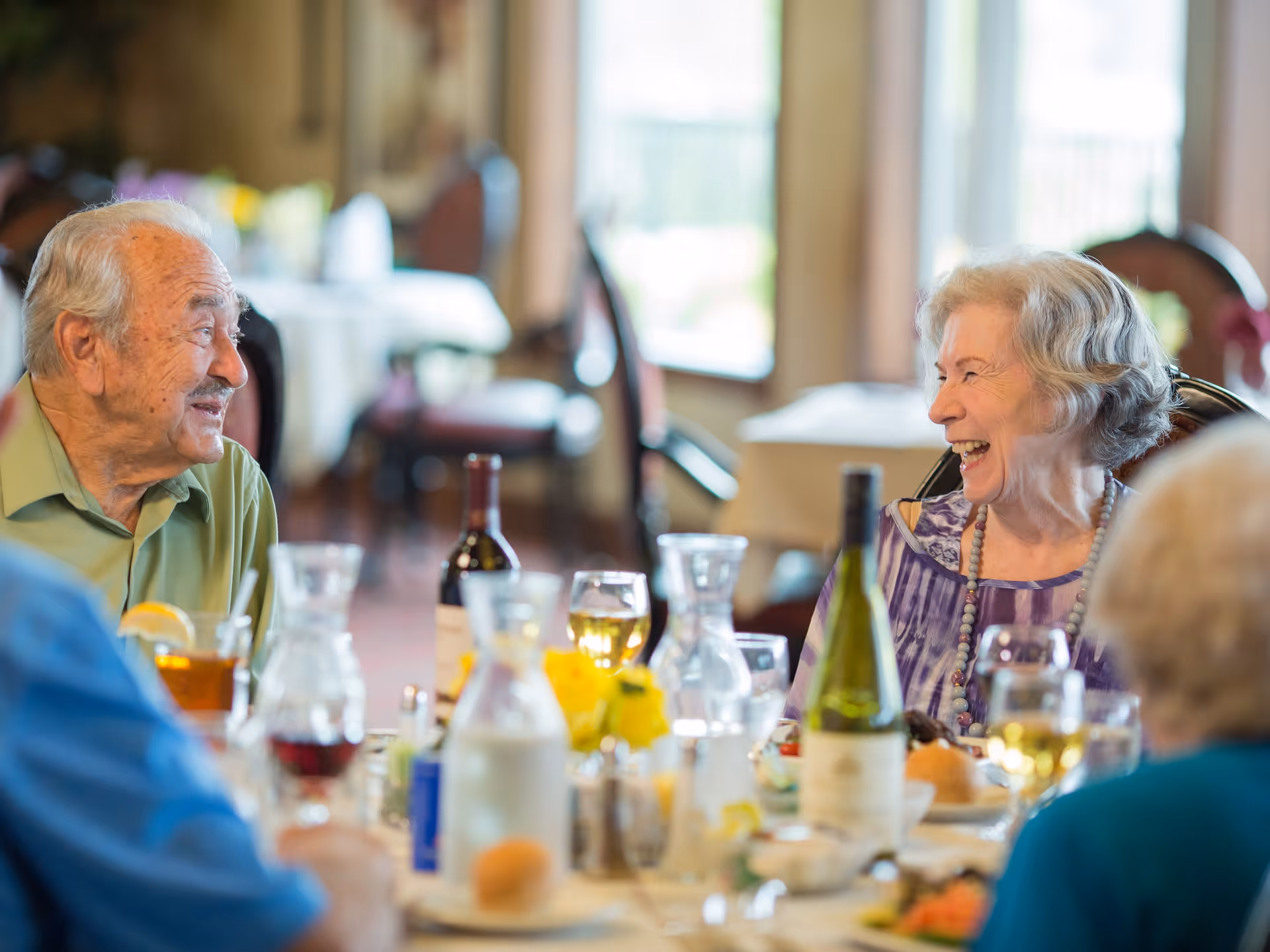 A group of elderly people sitting around a dining table in a well-lit room, enjoying a meal and engaging in lively conversation. The table is set with wine bottles, glasses, plates of food, and a small vase with yellow flowers. The atmosphere appears warm and cheerful.