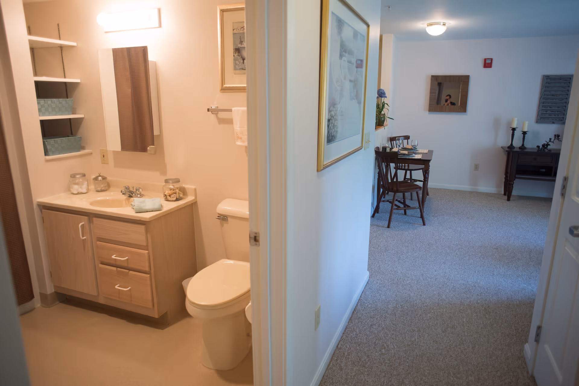 View of a small bathroom with a sink, mirror, and toilet on the left side, and a hallway leading to a dining area with a table and chairs on the right side. The bathroom has light wood cabinetry and shelves with storage baskets. The dining area has carpeted flooring and wall decorations.