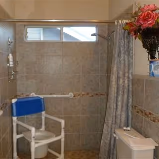 A bathroom with beige tiled walls and floor, featuring a shower area with a handheld showerhead, a white shower chair with a blue backrest, a grab bar on the wall, a shower curtain, and a toilet with a floral arrangement on a shelf above it.