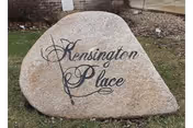 A large natural stone with the words 'Kensington Place' elegantly engraved on its surface, placed outdoors on a grassy area with some bushes and a sidewalk in the background.