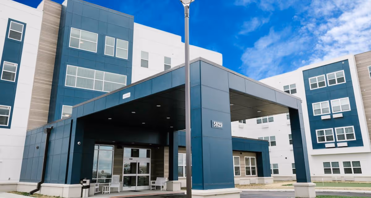 Exterior view of a modern multi-story senior living facility building with blue and white panels, large windows, and a covered entrance area with benches. The sky is partly cloudy and blue.