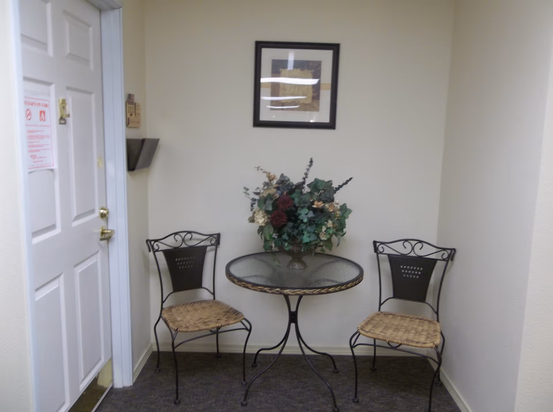 Small seating area with two metal chairs with woven seats and a round glass-top table with a floral arrangement on it, placed against a beige wall with a framed picture above. A white door with a notice is visible on the left side.