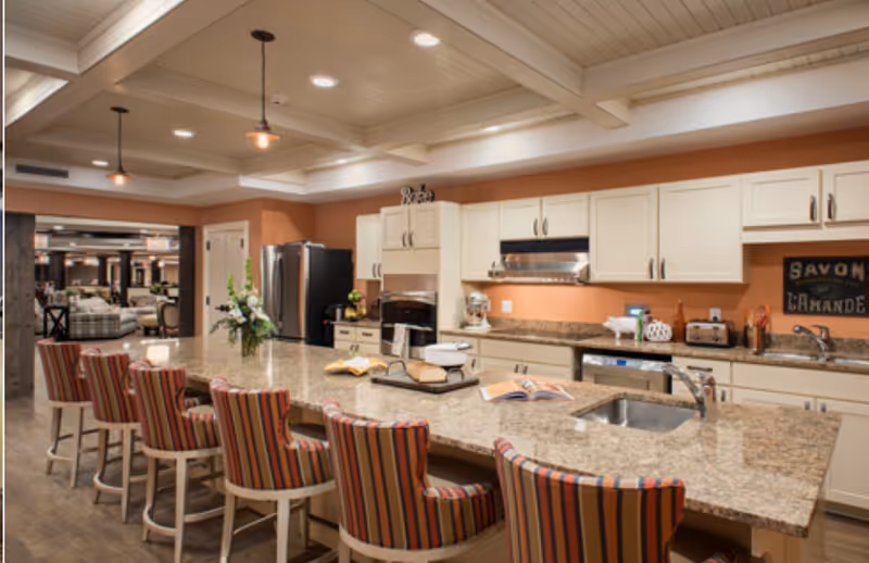 Spacious kitchen with a long granite island lined with striped bar stools, cream cabinets, and stainless appliances opening to a seating area.