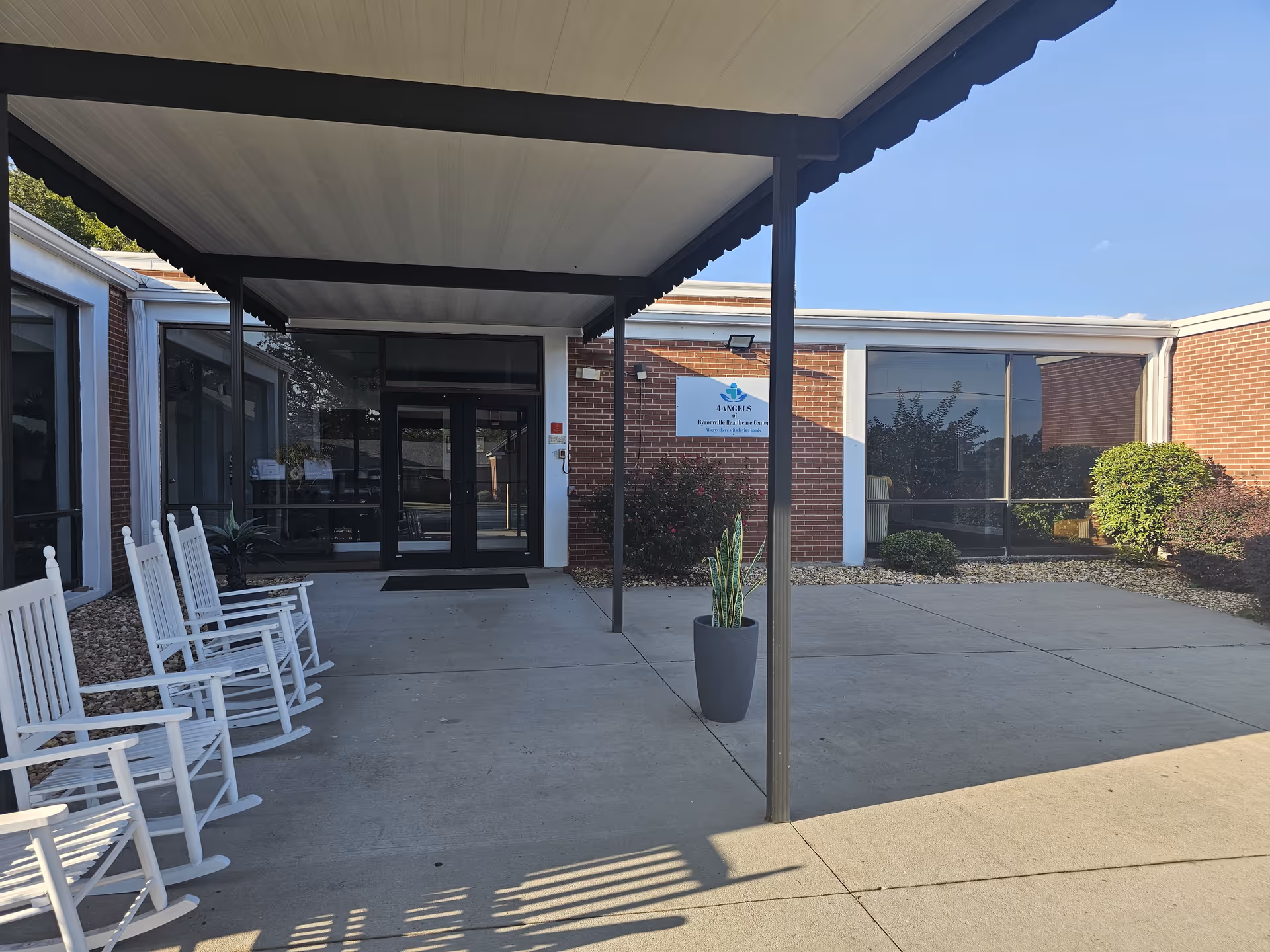 Covered entrance and driveway of a brick senior living facility with white rocking chairs, a potted plant, and a sign by the door.