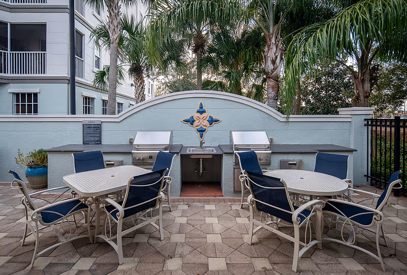 Outdoor patio area with two round tables surrounded by blue cushioned chairs. Behind the tables are two stainless steel grills and a sink set into a light blue wall with a decorative tile design. Palm trees and part of a building are visible in the background.