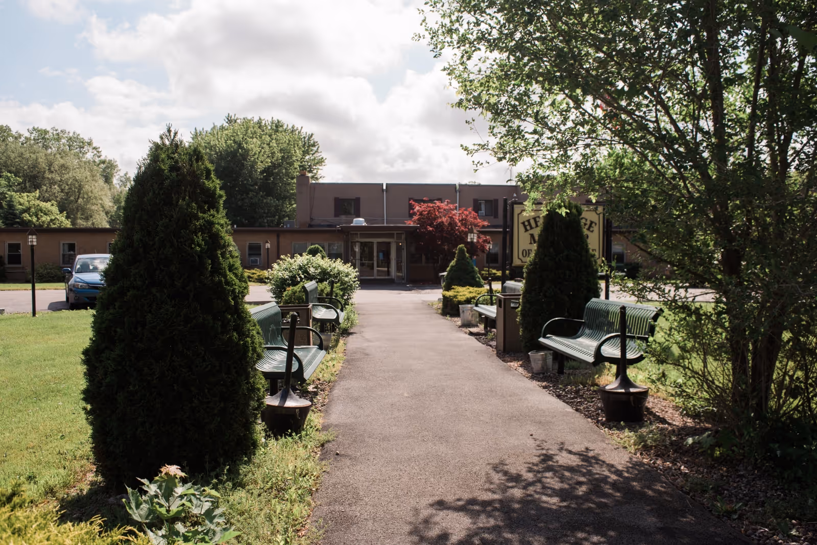 A paved walkway leading to the entrance of a single-story building with benches and trimmed bushes on either side. The area is surrounded by green grass and trees under a partly cloudy sky.