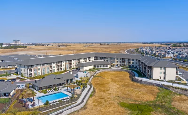 Aerial view of Ansel Park Independent Living facility showing a large, U-shaped three-story building surrounding an outdoor swimming pool area with lounge chairs and pergolas. The facility is situated next to open dry grassland and a nearby residential neighborhood with a clear blue sky above.