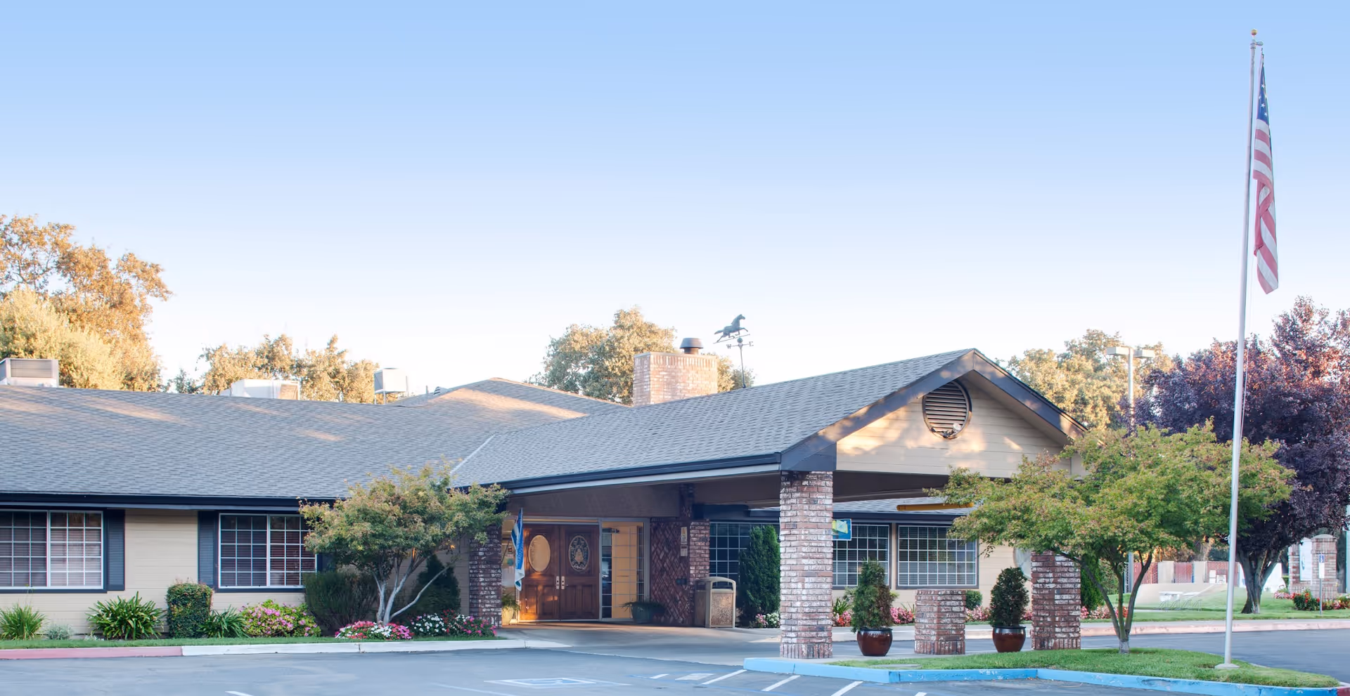Front exterior view of Creekside Care Center, a single-story building with a covered entrance supported by brick columns, surrounded by trees and landscaping, with an American flag on a flagpole to the right.