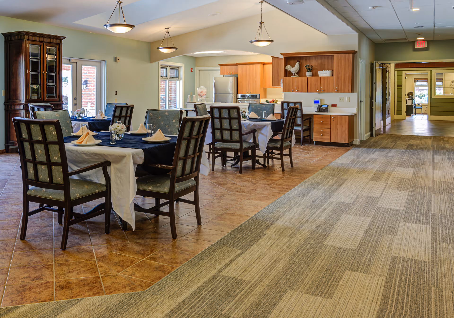 A dining area in a senior living facility with tables covered in white and navy blue tablecloths, set with napkins and glassware. The room has wooden chairs with patterned upholstery, tile flooring in the dining section, and carpet in the adjacent hallway. In the background, there is a kitchen area with wooden cabinets, a stainless steel refrigerator, and decorative items on the shelves. The space is well-lit with hanging light fixtures and natural light from windows and a glass door.