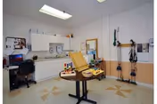 A therapy or rehabilitation room with a round table in the center holding various therapy tools and equipment. The room has white cabinets and countertops along one wall, a desk with a computer and chair, and exercise bands and other therapy devices mounted on the opposite wall. The floor has a patterned design, and the ceiling has fluorescent lighting.