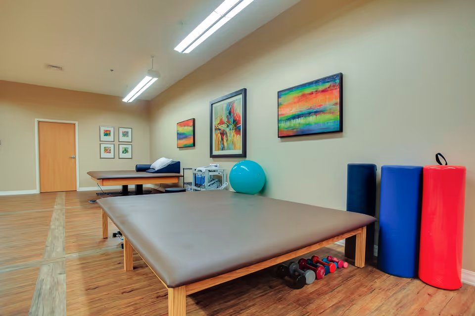 A therapy room with two padded treatment tables, colorful abstract paintings on the wall, a blue exercise ball, foam rollers in blue and red, and a set of small dumbbells on a wooden floor.