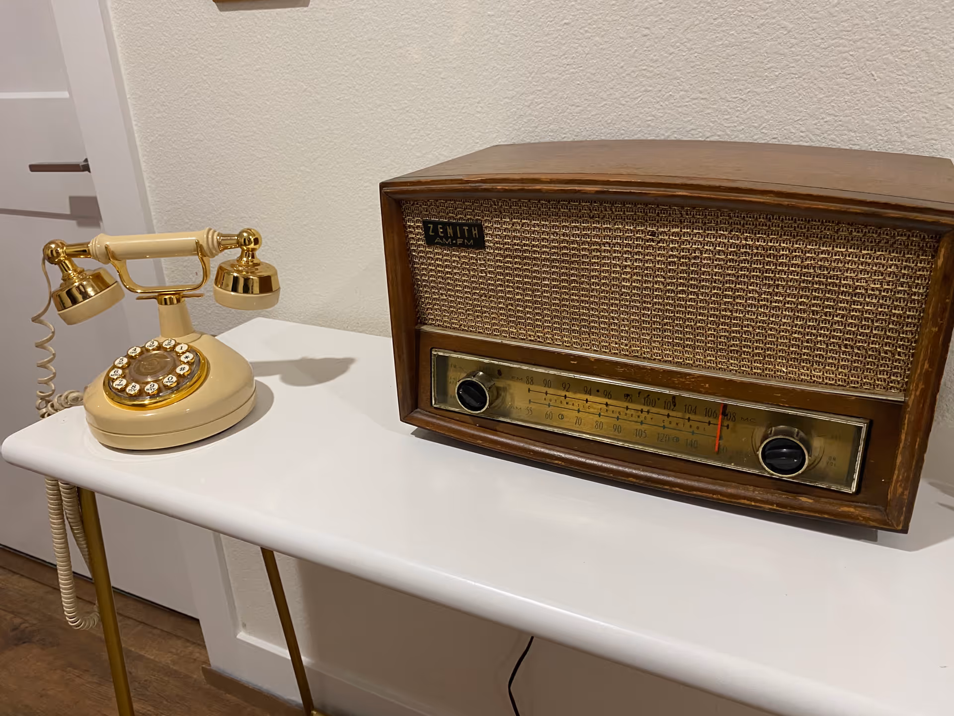 A vintage rotary dial telephone and a wooden Zenith AM-FM radio placed on a white table against a white wall.