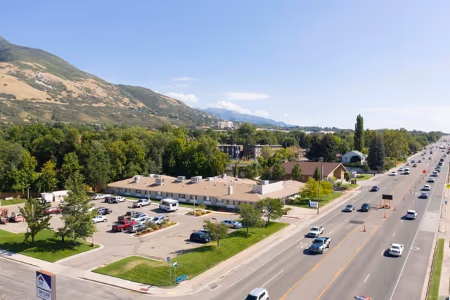 Aerial view of Harrison Pointe Healthcare And Rehabilitation facility surrounded by trees and greenery, with a parking lot in front and a busy road running alongside it. Mountains are visible in the background under a clear blue sky.