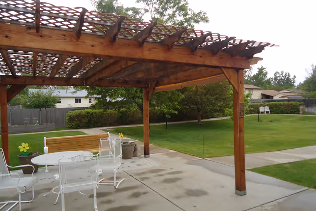 Outdoor patio area with a wooden pergola providing partial shade over a concrete surface. White metal chairs and a round table are arranged under the pergola. There is a wooden bench and potted plants nearby. The surrounding area features green grass, trees, and a paved walkway with residential houses and a wooden fence in the background.