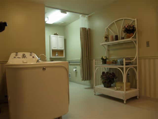 Interior view of a bathroom in Seminole Shores Living Center featuring a walk-in bathtub on the left, a white wicker shelf unit with decorative items on the right, and a small cabinet and toilet paper holder visible in the background.