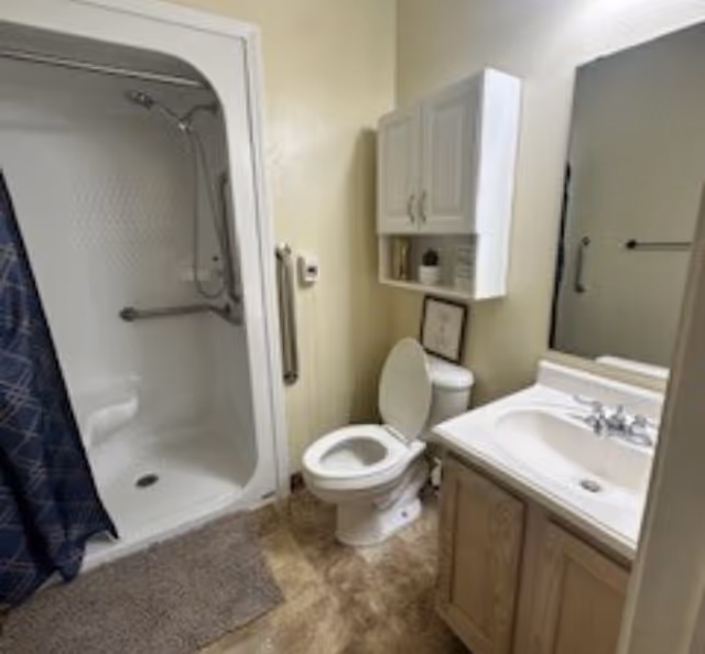 A bathroom with a white toilet, a sink with a wooden cabinet, a large mirror above the sink, a white wall-mounted cabinet above the toilet, and a shower with a blue patterned curtain and a built-in seat.