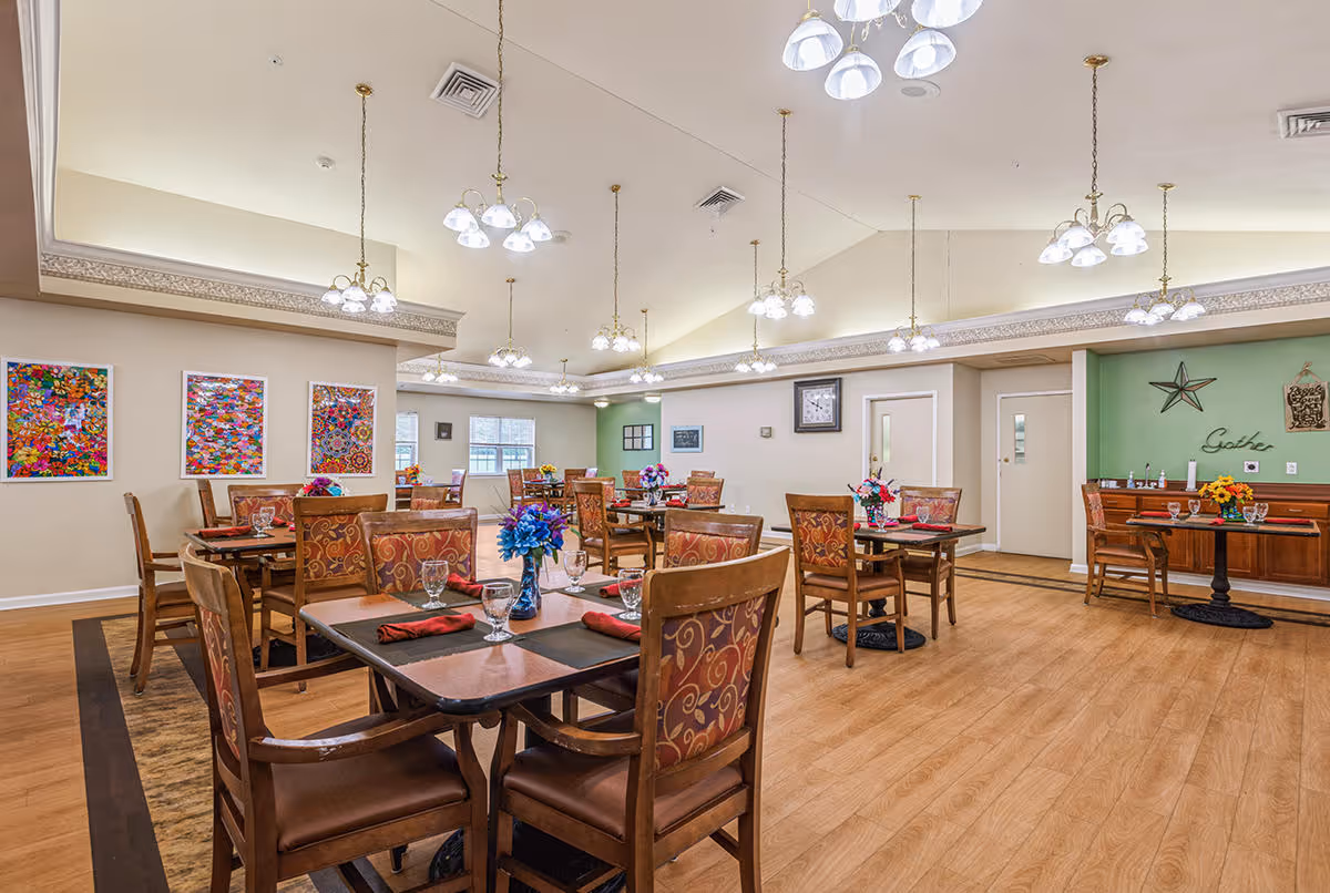 A spacious dining room with multiple wooden tables and chairs arranged neatly. Each table is set with placemats, glasses, and napkins, and decorated with colorful flower centerpieces. The room features a high ceiling with multiple hanging light fixtures, light-colored walls with colorful artwork, and a green accent wall with decorative items including a star and the word 'Gather'.