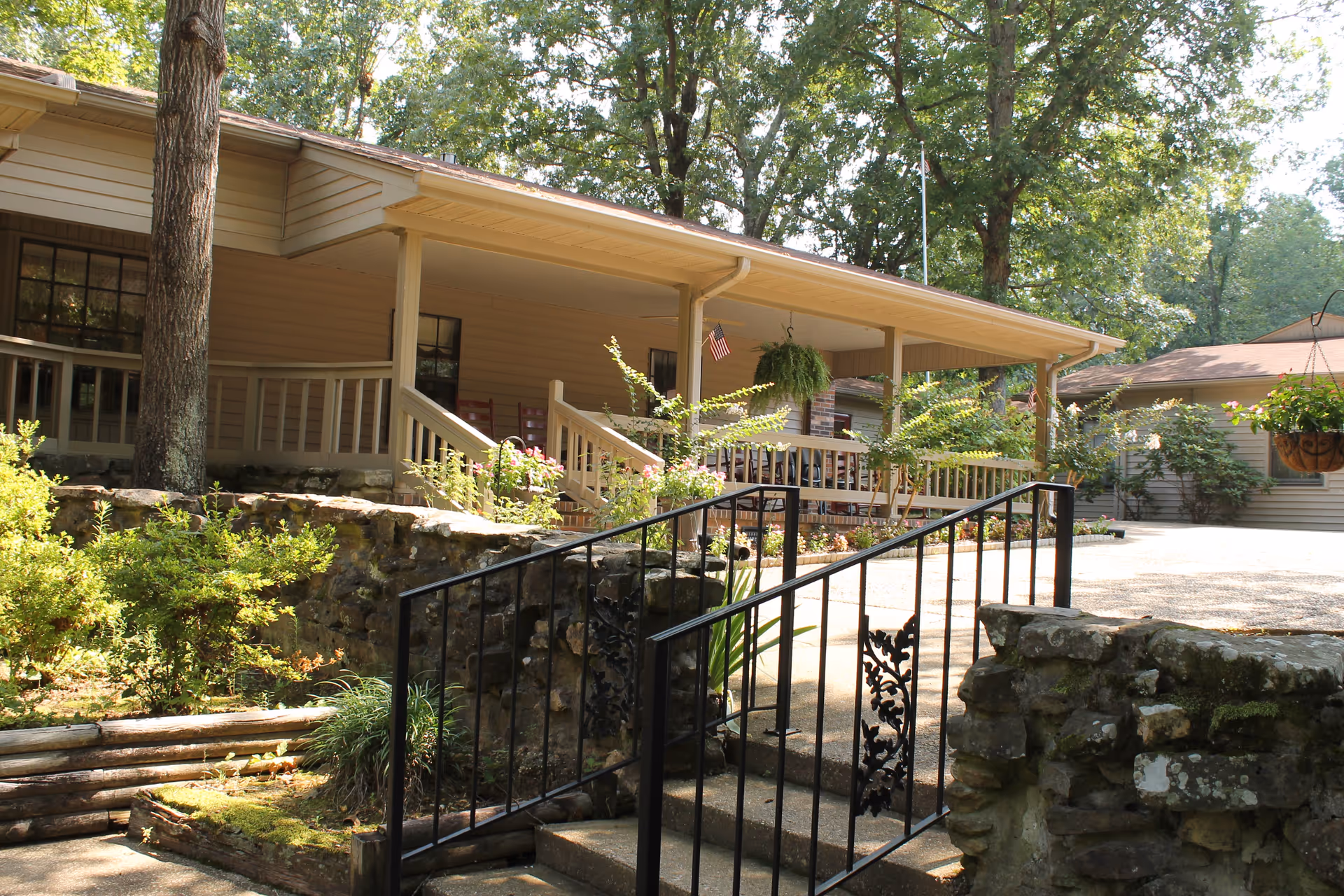 A single-story building with a covered porch surrounded by trees and greenery. The porch has railings and several chairs, with hanging plants and an American flag. Stone steps with a black metal railing lead up to the porch area.