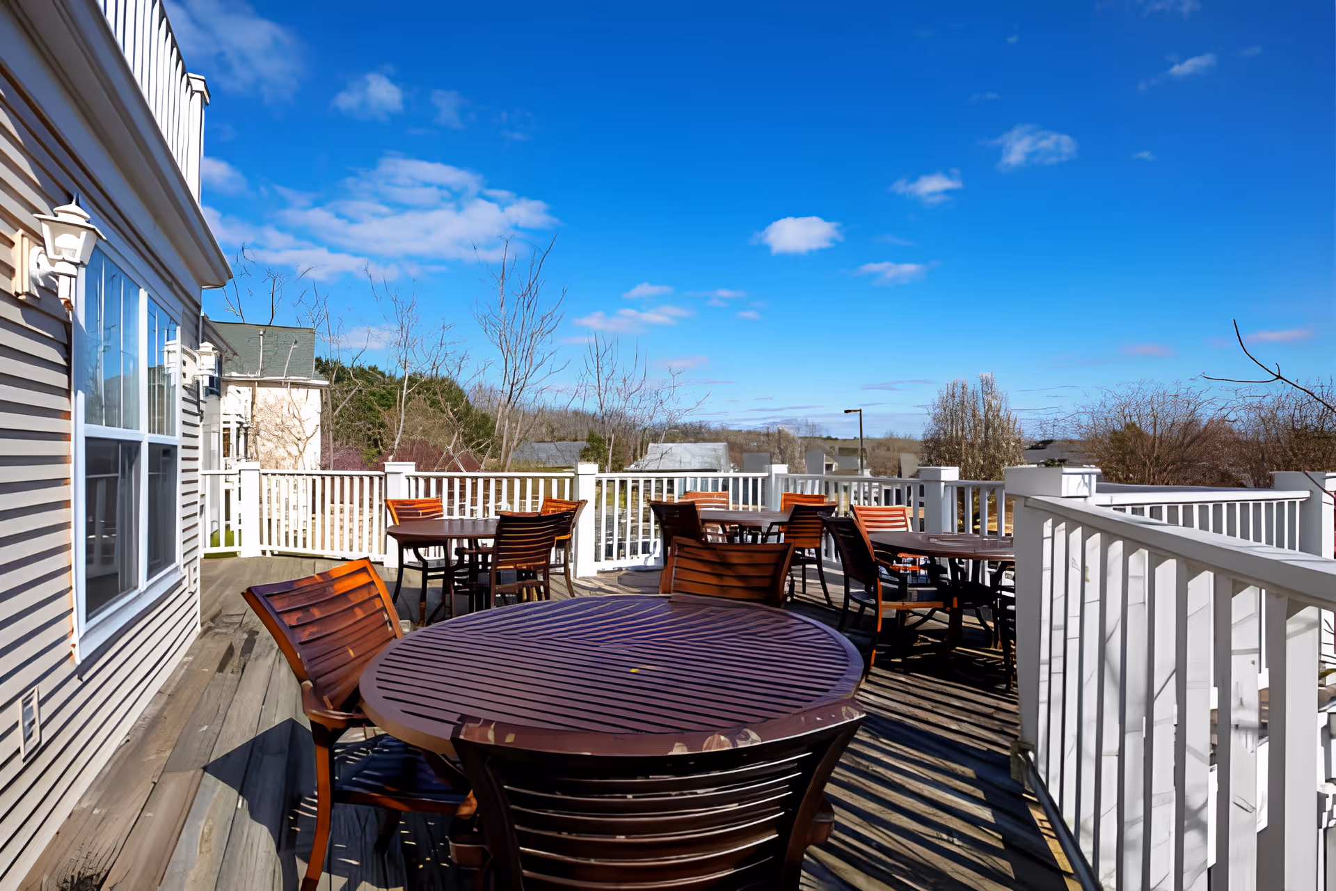Outdoor deck area with multiple round wooden tables and chairs, white railing surrounding the deck, adjacent to a building with beige siding and windows, under a clear blue sky with some clouds.