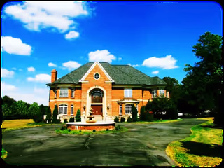Large two-story brick house with a steep roof and multiple windows, surrounded by trees and greenery under a blue sky with scattered clouds. A circular driveway with a small fountain in the center is in front of the house.