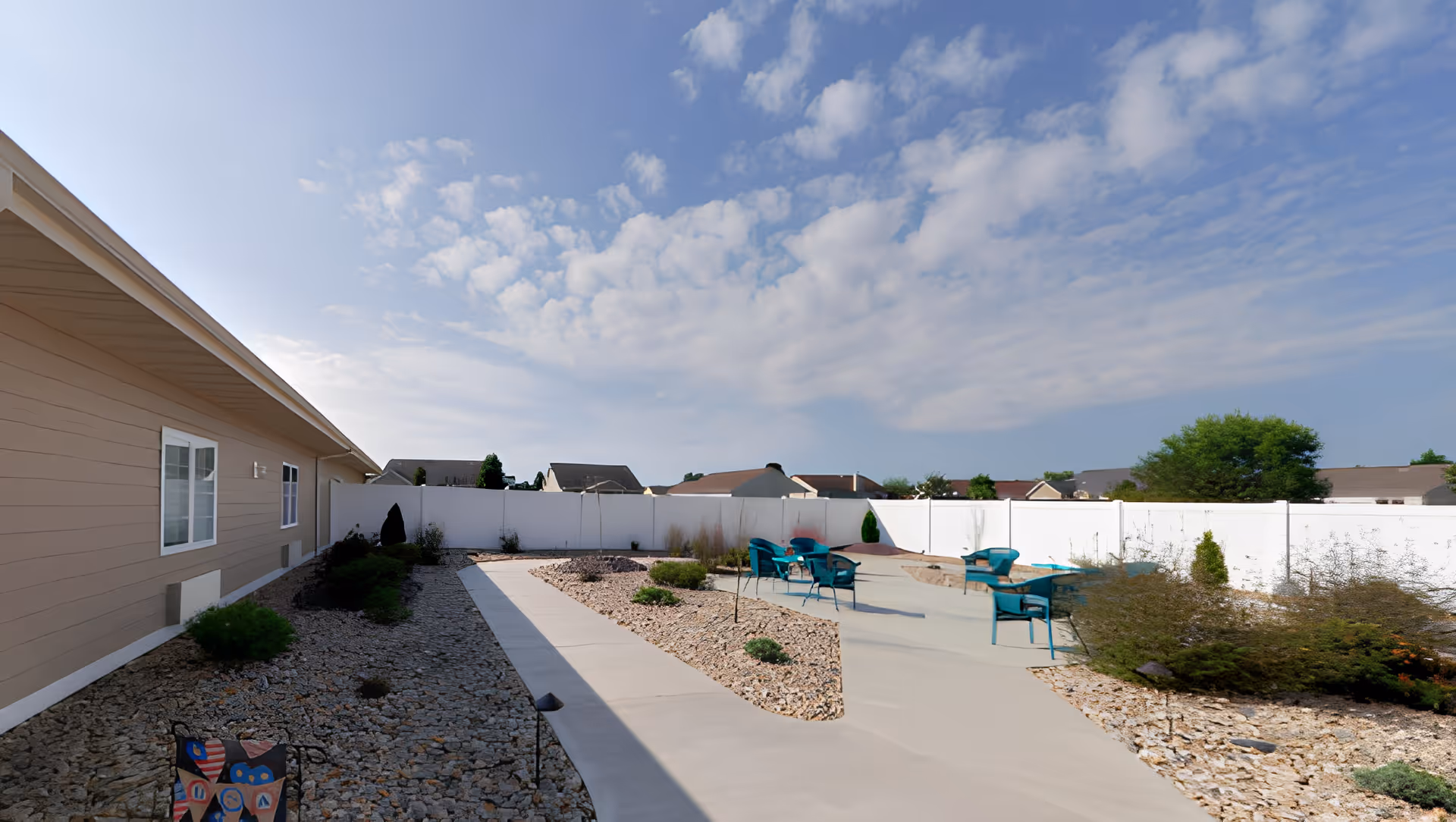 Outdoor patio area at Brightly Senior Living with a concrete walkway, several teal chairs arranged in small groups, surrounded by landscaping with rocks and shrubs, under a partly cloudy blue sky.