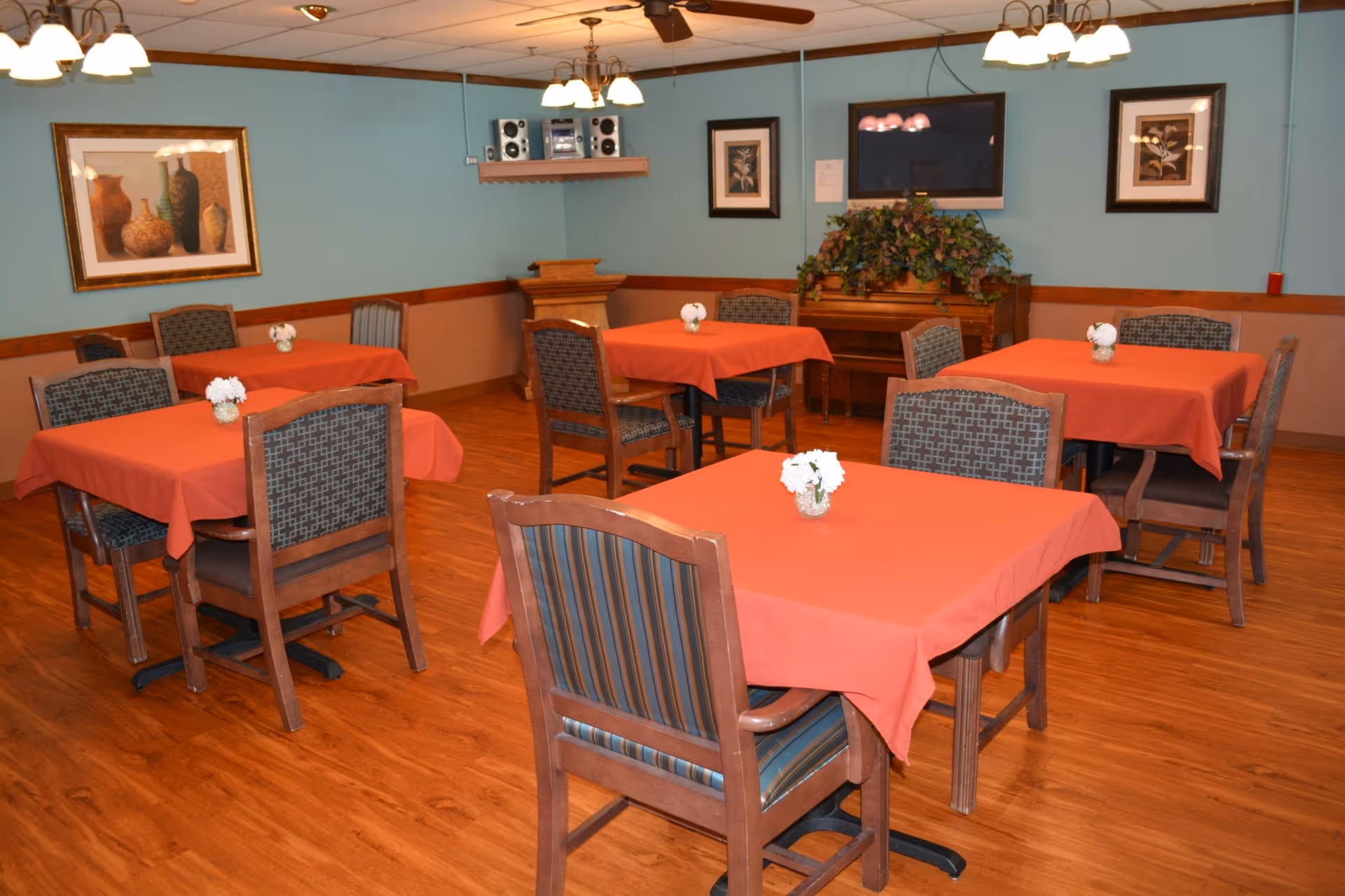 Dining room with multiple tables covered in red tablecloths and wooden chairs and small floral centerpieces.