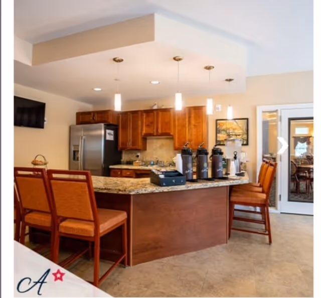 Open kitchen area with a granite island, bar stools, coffee dispensers and a stainless refrigerator in a senior living common space.