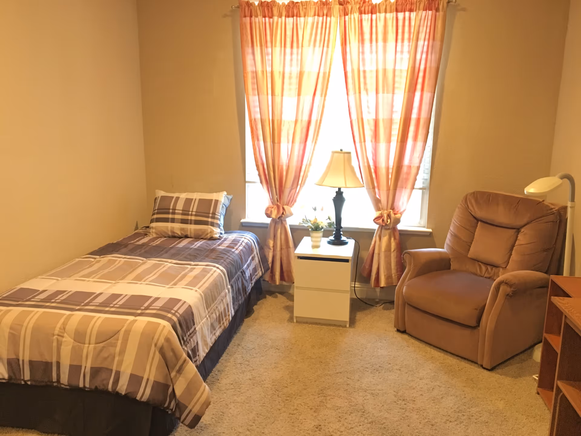 A cozy bedroom with a single bed covered in a plaid comforter and matching pillow. Next to the bed is a white nightstand with a black lamp and a small potted plant. The window behind the nightstand has orange and white checkered curtains tied in the middle. To the right of the nightstand is a comfortable brown armchair with a small pillow and a white floor lamp behind it. The room has beige walls and carpeted floor.